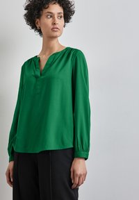 Blusa verde realizzata in tessuto morbido, con scollatura a V, maniche lunghe con polsini arricciati e una vestibilità rilassata. Abbinata a pantaloni neri.