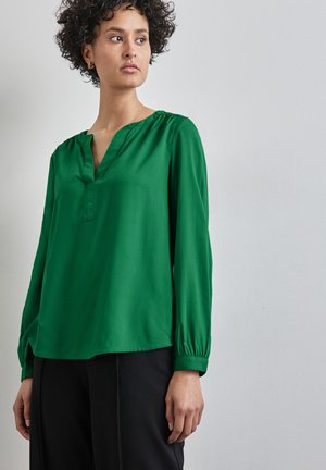 Green blouse made of smooth fabric, featuring a V-neck, long sleeves with gathered cuffs, and a relaxed fit. Styled with black pants.