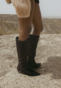 Botas marrones hasta la rodilla hechas de cuero texturizado con punta afilada y cremallera lateral. La superficie muestra un brillo sutil y una ligera textura similar al pelo.