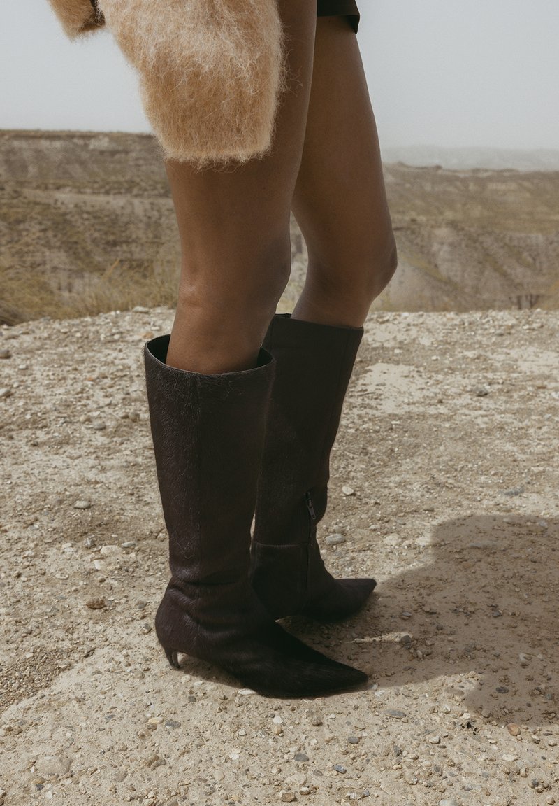 Botas marrones hasta la rodilla hechas de cuero texturizado con punta afilada y cremallera lateral. La superficie muestra un brillo sutil y una ligera textura similar al pelo.