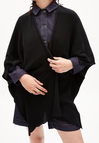 Svart stickad poncho med avspänd passform, med en öppen framsida, ribbad textur och scallopade kanter. Bärs över en denimskjorta.
