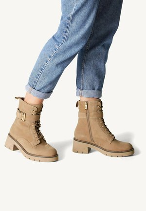 Bottines en suède beige avec un devant lacé, une fermeture éclair sur le côté, des détails en boucle et une semelle épaisse et texturée. Portées avec un jean bleu retroussé.