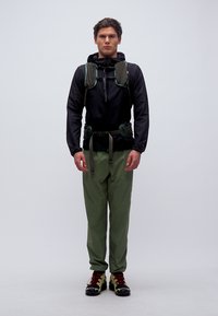 Άνδρας με μαύρο μπουφάν, πράσινο παντελόνι πεζοπορίας, σακίδιο πεζοπορίας και παπούτσια για τρέξιμο σε μονοπάτι, στέκεται μπροστά σε απλό φόντο.