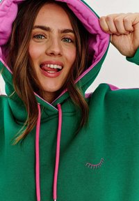 Zelená mikina s ružovým vnútorným podšívkou a šnúrkami. Na prednej strane sa nachádza malé vyšívané očko. Mäkká textúra látky.