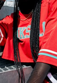 Rotes, kurzes Jersey mit türkisfarbener Schrift und Streifen, strukturierten Stoff. Lange, geflochtene Haare und geschichtete Outfits mit schwarzem Mesh-Unterlayer.