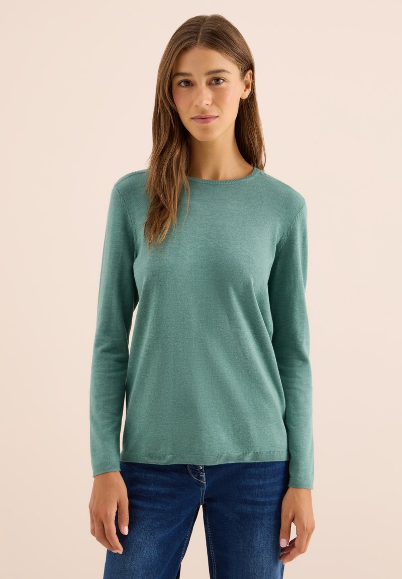 Jeune femme avec de longs cheveux bruns portant un pull à manches longues teal et un jean bleu, se tenant devant un fond beige uni.