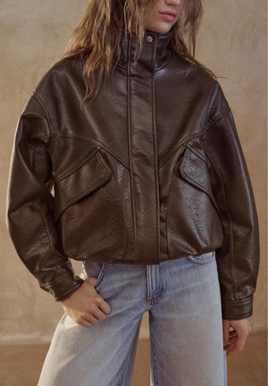 Frau trägt eine dunkelbraune Lederjacke mit großen Klappentaschen und hellblauen, hoch taillierten Jeans, steht mit einer Hand in der Tasche.