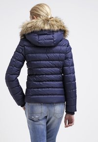 Žena viděná zezadu, oblečená v námorně modré prošívané bundě s kapucí lemovanou kožešinou a světlých modrých vybledlých džínách.