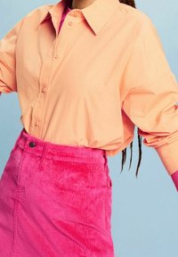 Personne portant une chemise pêche boutonnée rentrée dans une jupe en velours côtelé rose vif taille haute sur un fond bleu clair.
