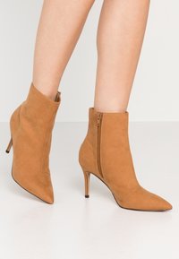 Bottes cheville en daim beige à bout pointu avec fermeture éclair latérale et talons aiguilles portées sur des jambes nues sur fond blanc.
