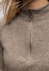 Grå ribbad polotröja med dragkedja framtill. Har en silverfärgad dragkedjeförslutning och en hög krage, tillverkad av texturerat stickat tyg.