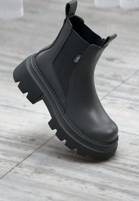 Svarta Chelsea-stövlar med en slät läderutsida, ribbade elastiska paneler och kraftiga, texturerade gummisulor. Har dragflikar för enkel påklädning.