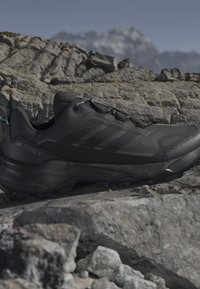 Scarpa da trekking nera su superficie rocciosa con la cima della montagna sfocata sullo sfondo sotto un cielo limpido.