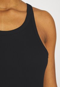 Gros plan sur l'épaule et la clavicule d'une personne portant un haut noir sans manches à côtes et un fin collier en or.