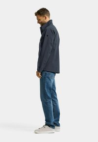 Marineblauw jack met een hoge kraag en steekzakken aan de voorkant, gecombineerd met blauwe jeans en lichtgrijze sneakers. Soepele textuur, casual pasvorm.