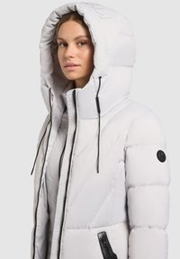 Polstret hvid jakke med stor hætte, prydet med sorte lynlåse og en sidelomme. Tekstureret materiale med diagonalt quiltet mønster.