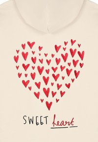 Sudadera cremosa con un diseño de corazón formado por varios corazones rojos, con el texto "SWEET heart" en negro y acentos en rojo. Textura suave.