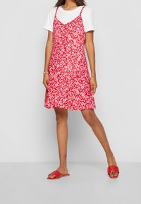 Röd blommig klänning med justerbara spaghetti-remmar, över en vit t-shirt. Kombinerad med röda platta sandaler. Slätt tyg, avslappnad passform.