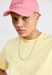 Casquette rose avec logo brodé, t-shirt jaune avec marque en relief, collier en chaîne argenté et tatouage en forme de cœur sur le cou.