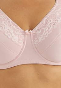 Weicher rosa BH mit Spitzenbesatz und kleinem Schleifendetail, getragen auf nackter Haut, Nahaufnahme des Brustbereichs.