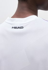 Κοντινή λήψη του λαιμού και του άνω μέρους της πλάτης ενός ατόμου που φοράει λευκό διάτρητο αθλητικό πουκάμισο με μαύρο λογότυπο "HEAD" κοντά στο γιακά.