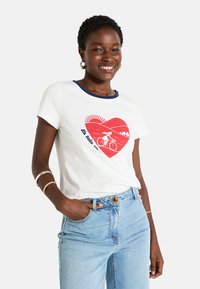 Camiseta blanca de algodón con un cuello acanalado azul marino, que presenta un gráfico de un corazón rojo con un ciclista y un sol, combinada con unos jeans de denim azul.
