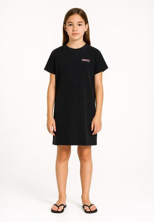 Junges Mädchen steht aufrecht, trägt ein schwarzes kurzärmliges Kleid mit dem Schriftzug "PROTECT" und schwarze Flip-Flops vor weißem Hintergrund.