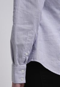 Randig skjorta med blå och vita vertikala ränder, långa ärmar, knäppning vid ärmsluten och en liten logotyp på sidan. Tyget ser slätt ut.