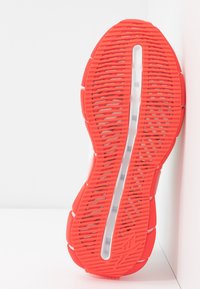 Semelle de chaussure en caoutchouc rouge vif avec des rainures et des crêtes texturées, présentant une zone centrale transparente. Des sections blanches délimitent les bords.