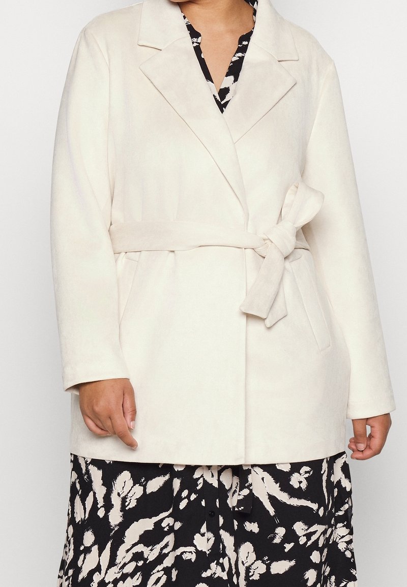 Femme portant un manteau ceinturé beige clair sur une robe à motifs noir et blanc, montrée du cou jusqu'au milieu de la cuisse.