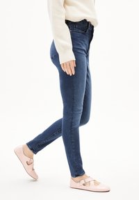 Tummansinisiä kapealahkeisia farkkuja yhdistettynä vaaleanpunaisiin ballerinoihin, joissa on leikkauskoristeluja ja solki. Neuletakissa on teksturoitu, neulottu malli.