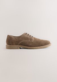 Chaussure Oxford homme en daim marron avec laçage à l'avant et semelle crêpe sur fond clair uni.