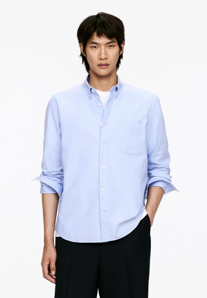 Hellblaues Hemd mit langen Ärmeln und Brusttasche, aus glattem Stoff gefertigt, mit Kragen und hochgerollten Ärmeln.