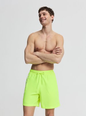 Giovane uomo a torso nudo sorridente con le braccia incrociate, indossa pantaloncini da bagno verdi neon luminosi, in piedi davanti a uno sfondo chiaro e semplice.