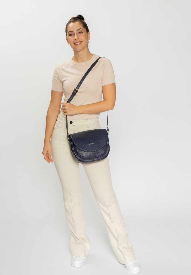 Bolso cruzado de cuero azul marino con solapa curva, superficie texturizada, correa ajustable y accesorios en tono dorado, llevado por un modelo sobre un fondo neutro.