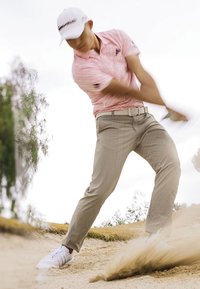 Golfista ubrany w różową koszulę i białą czapkę wykonujący uderzenie z piasku z bunkra, z piaskiem tryskającym w górę i rozmytymi drzewami w tle.