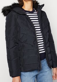 Padded schwarze Jacke mit Reißverschluss vorne, strukturiertem Stoff und Futter aus Kunstfell in der Kapuze. Verfügt über seitliche Taschen und horizontale Nähte.