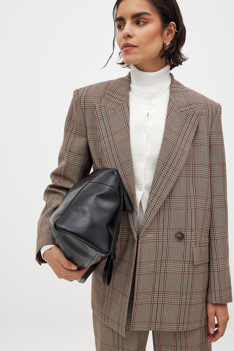 Geruite blazer in bruin en beige met rode accenten, gecombineerd met een crèmekleurige coltrui. Zwarte leren tas met een zachte textuur.