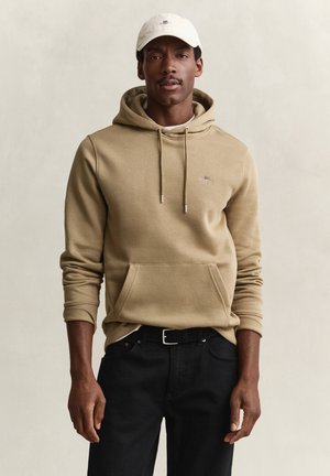 Man met een beige hoodie met voorzak, zwarte riem en broek, en een witte pet, staand tegen een effen achtergrond.