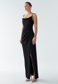 Black satin evening gown with cowl neckline, thin straps, side slit, and draping detail. Paired with gold accessories and high heels.