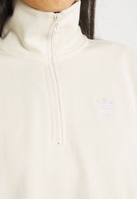 Krämfärgad zip-up sweatshirt i mjukt tyg, med hög krage och en liten, ljuslila Adidas-logotyp på vänster bröst.