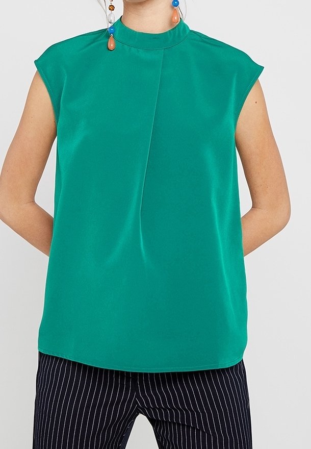 Femme portant un top sans manches bleu sarcelle à col montant avec un pli central, associé à un pantalon noir à fines rayures et des boucles d'oreilles pendantes colorées.