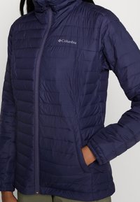 Kurtka puchowa Columbia w ciemnym granacie, z połączeniem gładkiego i teksturowanego materiału, pionowym pikowaniem, wysokim kołnierzem i kieszeniami na zamek.