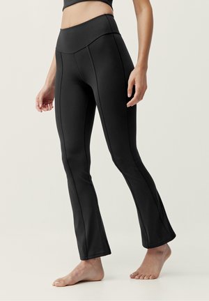 Kvinde iført sort højtaljede vide leggings, stående barfodet mod en ensfarvet lys baggrund.