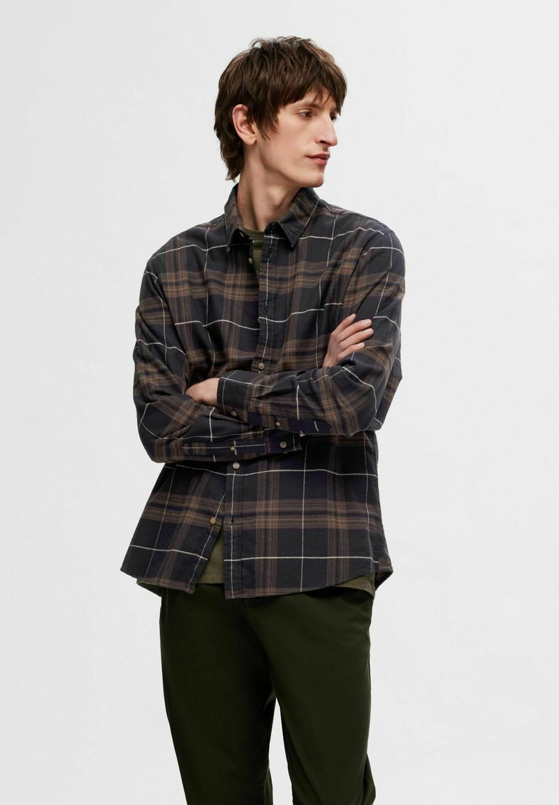 Camicia di flanella a scacchi in marrone scuro e blu navy, con righe a griglia bianche. Maniche lunghe, colletto abbottonato, fit rilassato, abbinata a pantaloni verde oliva.