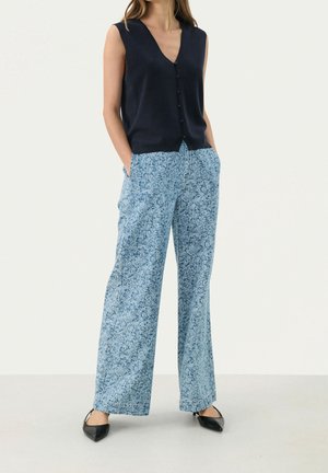 Femme portant un haut sans manches bleu marine boutonné, un pantalon large bleu clair à fleurs et des chaussures noires pointues, debout les mains dans les poches.