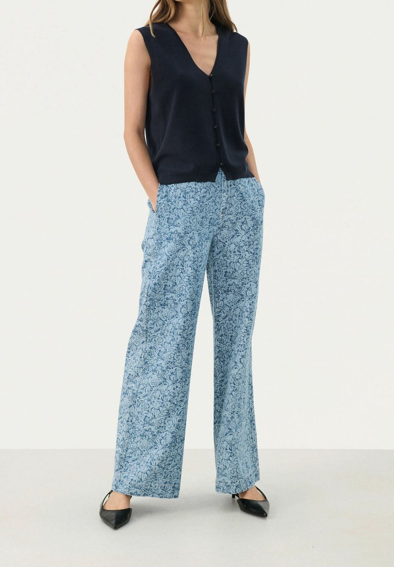 Femme portant un haut sans manches bleu marine boutonné, un pantalon large bleu clair à fleurs et des chaussures noires pointues, debout les mains dans les poches.