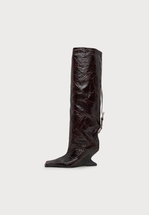 Une botte haute en cuir marron foncé, à la finition plissée, au bout pointu, au talon sculptural et à la boucle latérale, se détache sur un fond uni.