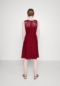 Burgundy spetsklänning med figurnära bodice och utsvängd kjol, med ett mönstrat spetsoverlägg och ärmlös design. Platta sandaler synliga.