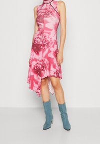 Różowa sukienka w kwiaty z wysokim dekoltem i asymetrycznym dołem, wykonana z elastycznego materiału. Połączona z jasnoniebieskimi botkami do kostek.
